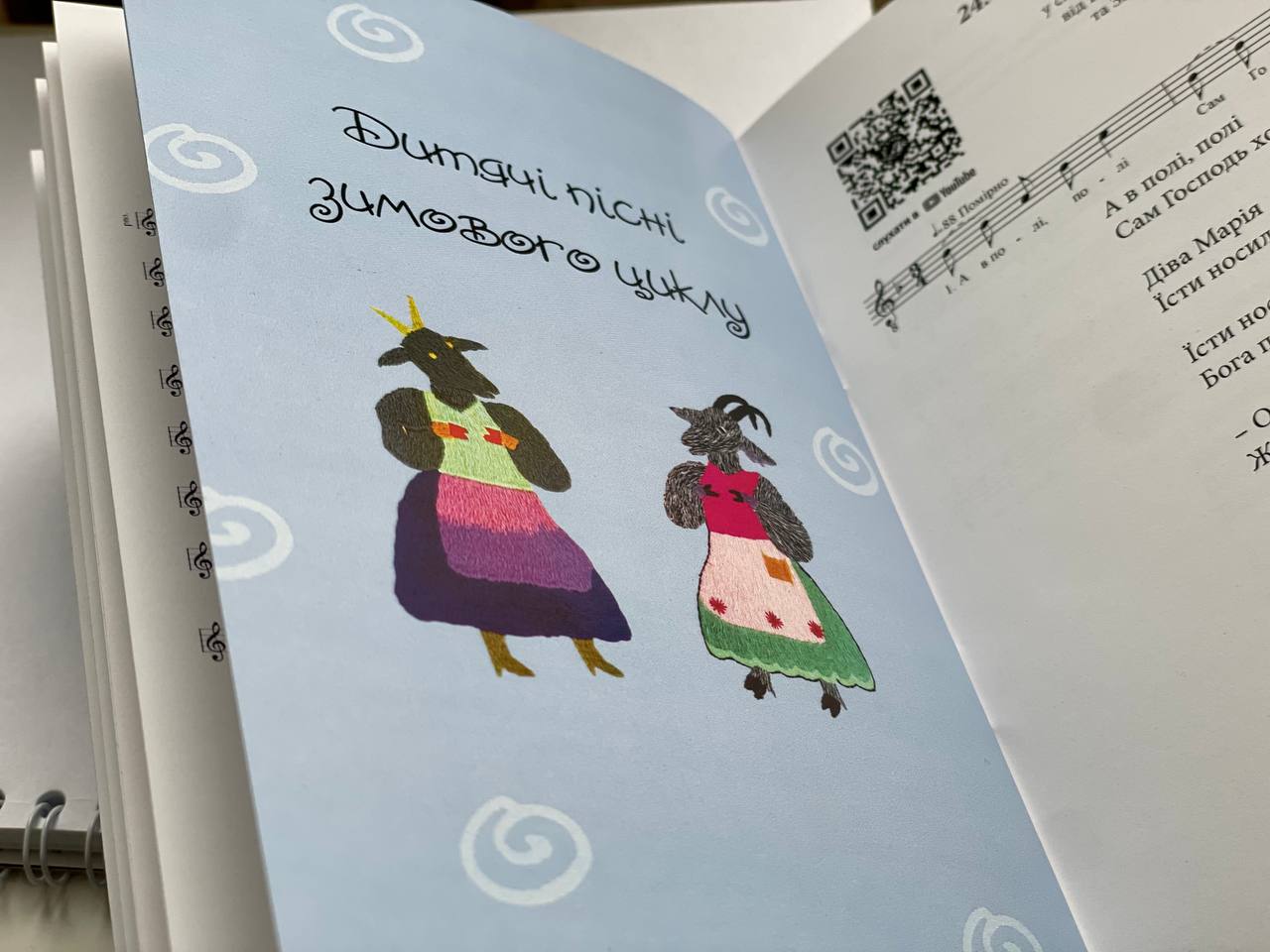 Розворот книги. На лівій сторінці на блакитному фоні міститься напис "Дитячі пісні зимового циклу" і ілюстрація двох антропоморфних фігур в яскравих костюмах. На правій сторінці видно ноти і текст пісні, а також QR-код у верхньому куті.